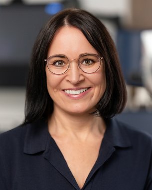 Eine Frau mit Brille lächelt freundlich in einem medizinischen Umfeld, mit einem optischen Gerät im Hintergrund.