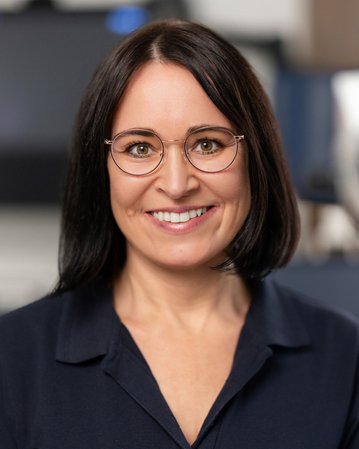 Eine Frau mit Brille lächelt freundlich in einem medizinischen Umfeld, mit einem optischen Gerät im Hintergrund.