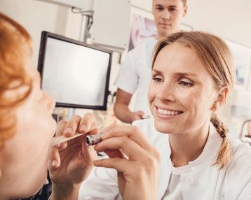 Eine Ärztin untersucht das Maul eines Patienten mit einem medizinischen Instrument, während ein weiterer medizinischer Fachmann im Hintergrund beobachtet. Die Umgebung wirkt klinisch und professionell.