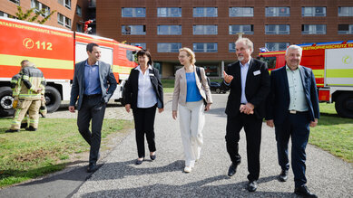 Fünf Personen gehen auf einem Weg, während Feuerwehrfahrzeuge im Hintergrund stehen. Sie scheinen an einer Veranstaltung oder Besichtigung teilzunehmen.