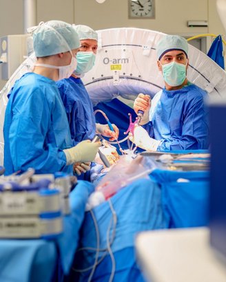 Drei medizinische Fachkräfte in blauen OP-Kitteln und Masken arbeiten an einem Patienten, während sie chirurgische Instrumente und Geräte verwenden. Im Hintergrund ist ein O-arm sichtbar, der für bildgebende Verfahren genutzt wird.
