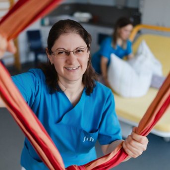 Eine Person in blauer Arbeitskleidung hält ein rotes und orangefarbenes Seil in der Hand, während im Hintergrund eine weitere Person auf einem Bett sitzt.