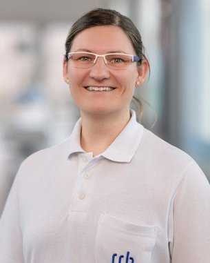 Eine lächelnde Frau mit Brille trägt ein weißes Poloshirt mit dem Logo "srh". Der Hintergrund ist unscharf und hell.