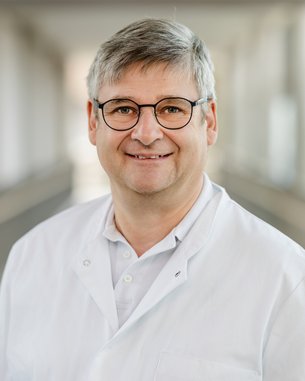 Ein Mann mit Brille und einem weißen Kittel lächelt in einem modernen Flur. Er hat graue Haare und trägt ein Polohemd darunter.