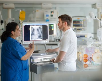 Eine Krankenschwester in blauer Uniform und ein Arzt im weißen Kittel besprechen eine Röntgenaufnahme der Lunge an einem Monitor. Der Raum ist mit medizinischen Geräten ausgestattet, die für die Patientenversorgung verwendet werden.