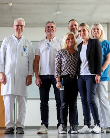 Sechs Personen, darunter ein Arzt in weißem Kittel, stehen lächelnd in einem modernen Innenraum. Sie tragen verschiedene Freizeitkleidung.