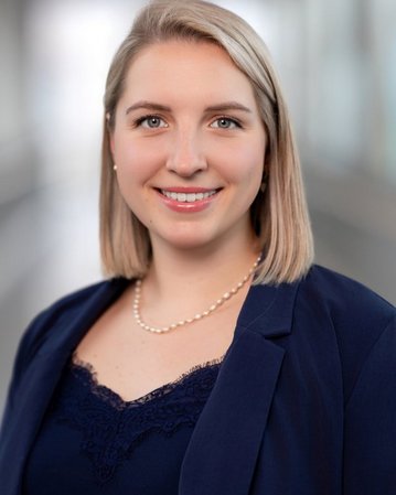 Eine junge Frau mit blonden, schulterlangen Haaren und einem blauen Blazer lächelt freundlich in die Kamera. Sie trägt eine Perlenkette.