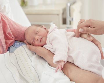 Ein Neugeborenes in einem rosa Overall schläft friedlich in den Händen einer Person, während eine andere Person sanft den Bauch des Babys streichelt.