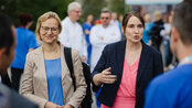 Zwei Frauen im Gespräch, eine in einem blauen Kleid und die andere in einem rosa Kleid, umgeben von Menschen in medizinischer Kleidung.