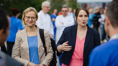 Zwei Frauen im Gespräch, eine in einem blauen Kleid und die andere in einem rosa Kleid, umgeben von Menschen in medizinischer Kleidung.