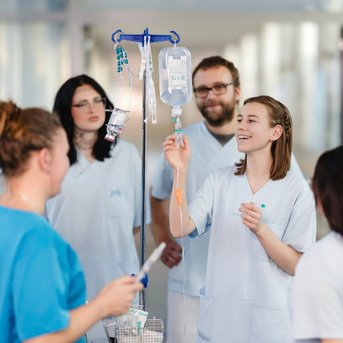 Eine Gruppe von Pflegekräften in weißen Kitteln steht um einen Infusionsständer und diskutiert die Anwendung einer Infusion.