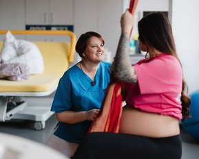 Eine schwangere Frau in einem pinken Oberteil interagiert mit einer Pflegekraft in blauer Uniform in einem Krankenhauszimmer.