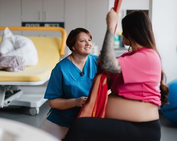 Eine schwangere Frau in einem pinken Oberteil interagiert mit einer Pflegekraft in blauer Uniform in einem Krankenhauszimmer.