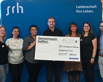 Gruppenszene mit sieben Personen, die einen Scheck über 15.000 € für die SRH Stiftung präsentieren. Hintergrund mit SRH-Logo.