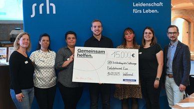 Gruppenszene mit sieben Personen, die einen Scheck über 15.000 € für die SRH Stiftung präsentieren. Hintergrund mit SRH-Logo.