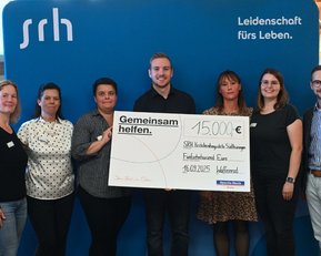 Gruppenszene mit sieben Personen, die einen Scheck über 15.000 € für die SRH Stiftung präsentieren. Hintergrund mit SRH-Logo.