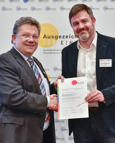 Zwei Männer schütteln sich die Hände, während einer ein Zertifikat hält. Im Hintergrund sind Logos und der Schriftzug „Ausgezeichnet für Kinder“ zu sehen.