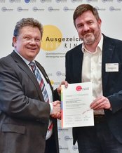 Zwei Männer schütteln sich die Hände, während einer ein Zertifikat hält. Im Hintergrund sind Logos und der Schriftzug „Ausgezeichnet für Kinder“ zu sehen.