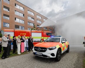 Ein Einsatzfahrzeug der Feuerwehr steht vor einem Gebäude, während Feuerwehrleute und Zuschauer applaudieren.