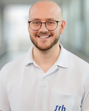 Ein junger Mann mit Glatze und Brille lächelt freundlich. Er trägt ein weißes Polohemd mit einem blauen Logo auf der Brust.