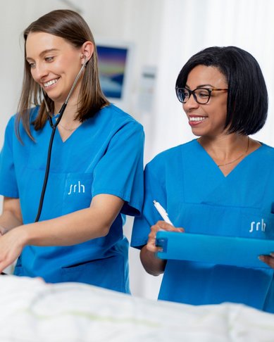 Zwei Krankenschwestern in blauen Uniformen stehen neben einem Patienten im Bett. Eine Schwester hält ein Stethoskop und lächelt, während die andere Notizen auf einem Clipboard macht. Der Patient sieht entspannt aus und lächelt zurück.
