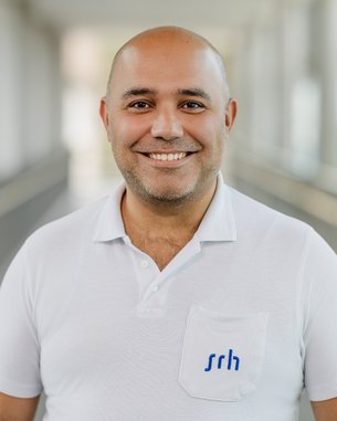 Ein lächelnder Mann mit Glatze trägt ein weißes Polohemd mit einem blauen Logo auf der Brust. Der Hintergrund ist unscharf.