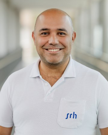 Ein lächelnder Mann mit Glatze trägt ein weißes Polohemd mit einem blauen Logo auf der Brust. Der Hintergrund ist unscharf.
