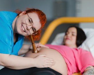 Eine schwangere Frau liegt auf einem Bett, während eine Hebamme mit einem Stethoskop ihren Bauch untersucht und lächelt.