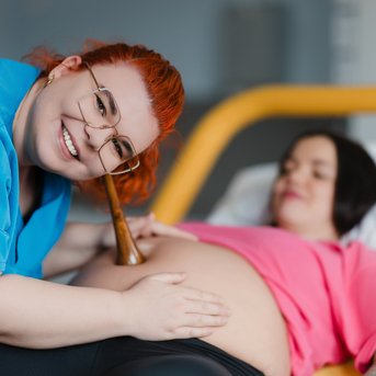 Eine schwangere Frau liegt auf einem Bett, während eine Hebamme mit einem Stethoskop ihren Bauch untersucht und lächelt.