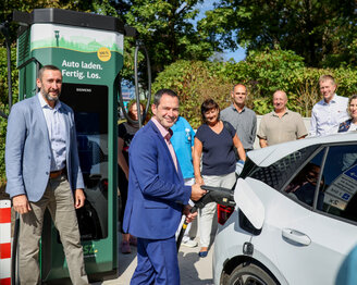 Eine Gruppe von Personen steht um eine Elektroauto-Ladestation, während ein Mann im Anzug das Auto auflädt.
