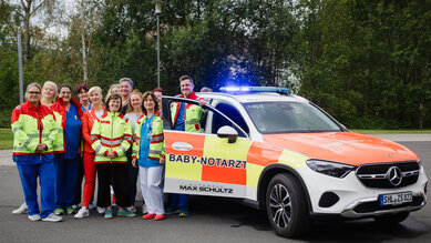 Eine Gruppe von Rettungskräften steht neben einem Einsatzfahrzeug mit der Aufschrift "BABY-NOTARZT". Das Fahrzeug hat ein blaues Blinklicht.