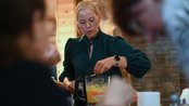 Eine Frau mit Brille und grünem Oberteil bereitet in einem Mixer eine Mischung vor, während andere Personen im Hintergrund sichtbar sind.