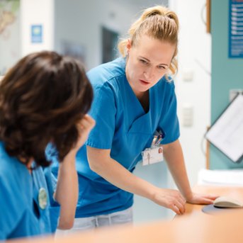 Zwei Krankenschwestern in blauen Uniformen unterhalten sich an einem Empfangstresen im Krankenhaus.