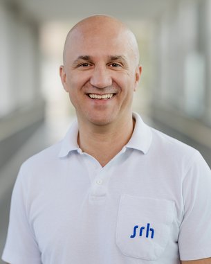 Lächelnder Mann mit Glatze trägt ein weißes Polohemd mit blauer Schrift "srh". Hintergrund ist unscharf und hell.