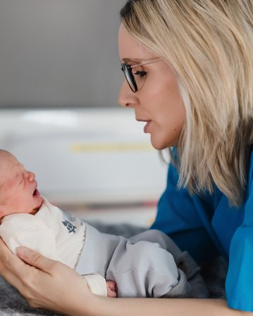 Eine Frau mit blonden Haaren und Brille hält ein weinendes Neugeborenes in ihren Händen und schaut es aufmerksam an.