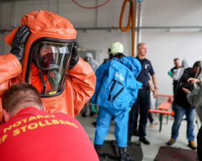 Person in orange Schutzanzug bereitet sich auf einen Einsatz vor, während andere Einsatzkräfte in Schutzkleidung im Hintergrund stehen.