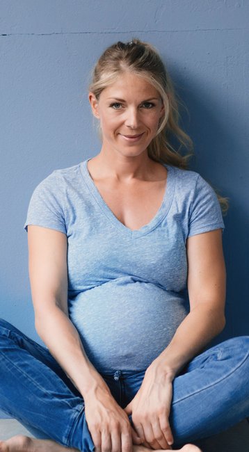 Eine schwangere Frau sitzt mit gekreuzten Beinen auf dem Boden vor einer blauen Wand. Sie trägt ein hellblaues T-Shirt und Jeans, und lächelt freundlich in die Kamera.