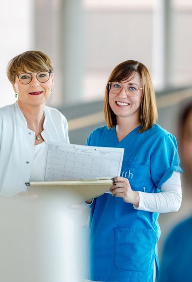 Zwei Frauen in medizinischer Kleidung stehen in einem modernen Krankenhausumfeld. Eine Frau hält ein Dokument, während die andere lächelt. Im Hintergrund ist eine weitere Person in blauer Kleidung unscharf zu sehen.