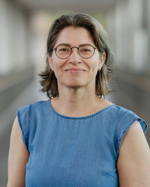 Frau mit kurzen, lockigen Haaren und Brille trägt ein ärmelloses Jeansoberteil. Sie lächelt freundlich in einem hellen, modernen Raum.