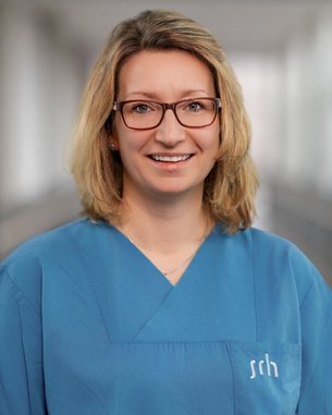 Eine Frau mit blonden Haaren trägt ein blaues medizinisches Outfit und Brille. Sie lächelt freundlich in die Kamera.