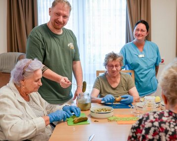 Eine Gruppe von vier Personen, darunter Senioren und ein Betreuer, bereitet gemeinsam Lebensmittel vor, während sie an einem Tisch sitzen.