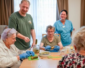 Eine Gruppe von vier Personen, darunter Senioren und ein Betreuer, bereitet gemeinsam Lebensmittel vor, während sie an einem Tisch sitzen.