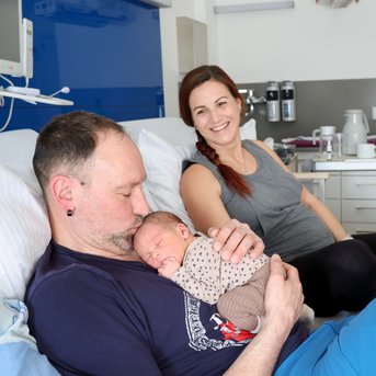 Ein Mann hält ein neugeborenes Baby in seinen Armen, während eine Frau lächelnd im Hintergrund sitzt. Sie befinden sich in einem Krankenhauszimmer.
