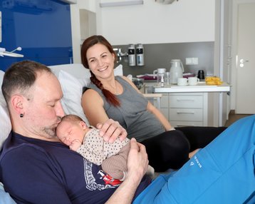 Ein Mann hält ein neugeborenes Baby in seinen Armen, während eine Frau lächelnd im Hintergrund sitzt. Sie befinden sich in einem Krankenhauszimmer.