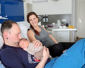 Ein Mann hält ein neugeborenes Baby in seinen Armen, während eine Frau lächelnd im Hintergrund sitzt. Sie befinden sich in einem Krankenhauszimmer.