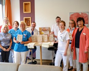 Eine Gruppe von neun Personen in Arbeitskleidung steht um einen Tisch mit mehreren Buchstaben und einem Foto. Sie lächeln in die Kamera.