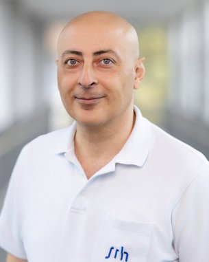 Mann mit Glatze trägt ein weißes Polohemd mit blauer Schrift. Er steht in einem hellen, modernen Innenraum.