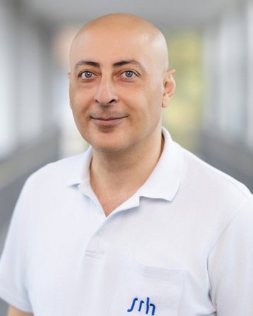 Mann mit Glatze trägt ein weißes Polohemd mit blauer Schrift. Er steht in einem hellen, modernen Innenraum.