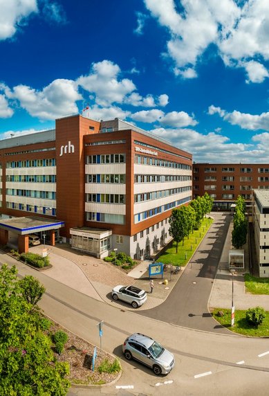 Moderne Gebäude der Fachhochschule mit grünen Flächen und Parkplatz, umgeben von Bäumen und einem klaren Himmel.