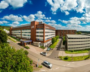 Moderne Gebäude der Fachhochschule mit grünen Flächen und Parkplatz, umgeben von Bäumen und einem klaren Himmel.